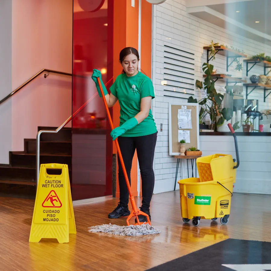 Janitor in green shirt mops wooden floor near caution wet floor sign and yellow cleaning bucket in bright lobby.