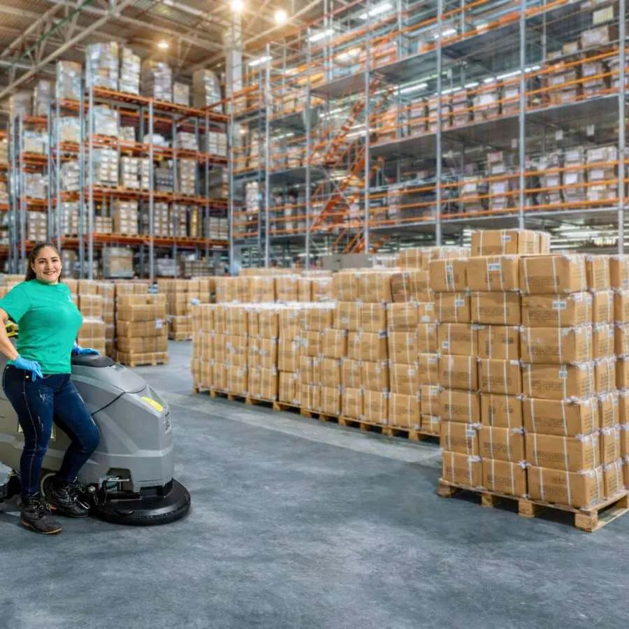 Warehouse worker with green shirt standing on floor cleaning machine amidst pallets of boxed goods in large warehouse.