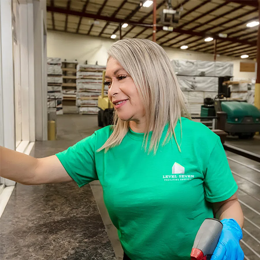 Woman in green Level Seven Facilities Services shirt cleaning a countertop with spray bottle in an industrial setting