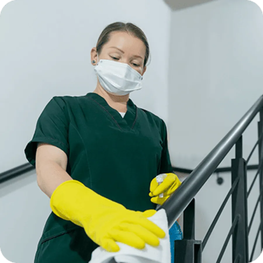 Woman in scrubs and yellow gloves disinfecting stair railing with spray and cloth indoors.
