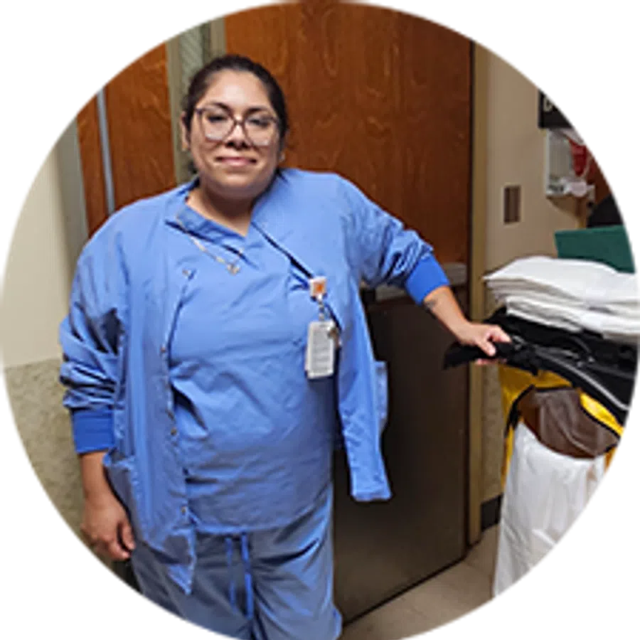 Smiling healthcare worker in blue scrubs and glasses standing in a hospital room next to medical supplies.