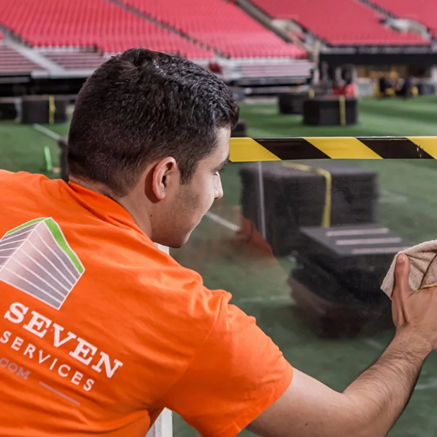 Worker in orange shirt cleaning glass barrier inside a large empty stadium with red seats and green field.