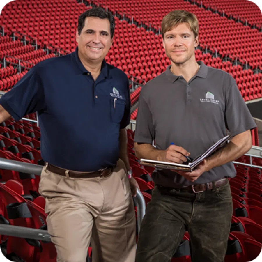 Two men in company shirts stand in a stadium with red seats, one holding a clipboard and pen.