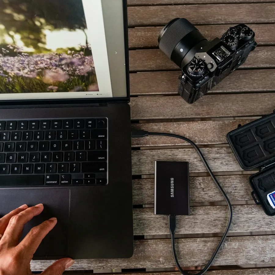 Hand using laptop next to a camera, external Samsung SSD, and SD card case on wooden table.