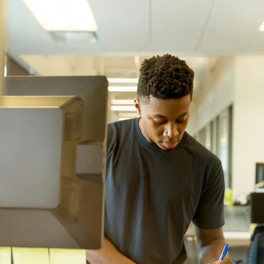 Young man standing at desk in office, writing notes in notebook with computer and keyboard nearby