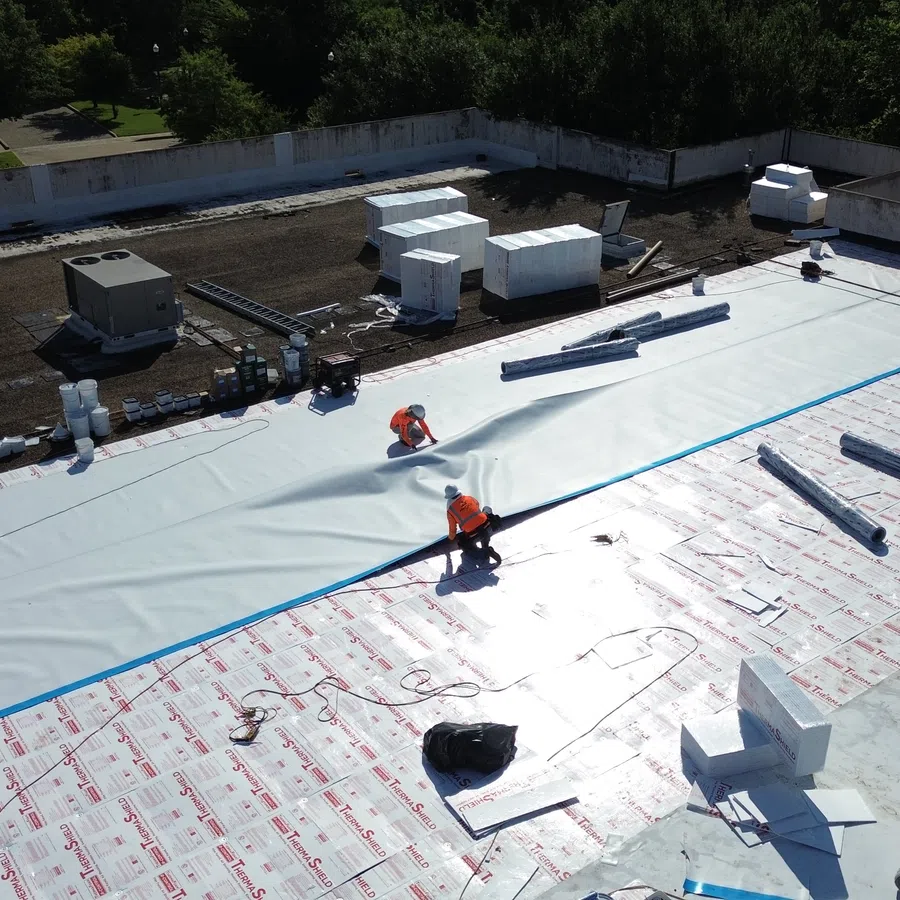 Aerial view of a Mattco crew installing a white TPO membrane roofing system on a commercial property in College Station, Texas, with workers in orange safety vests rolling out the membrane over ThermaShield insulation board on a large flat roof.