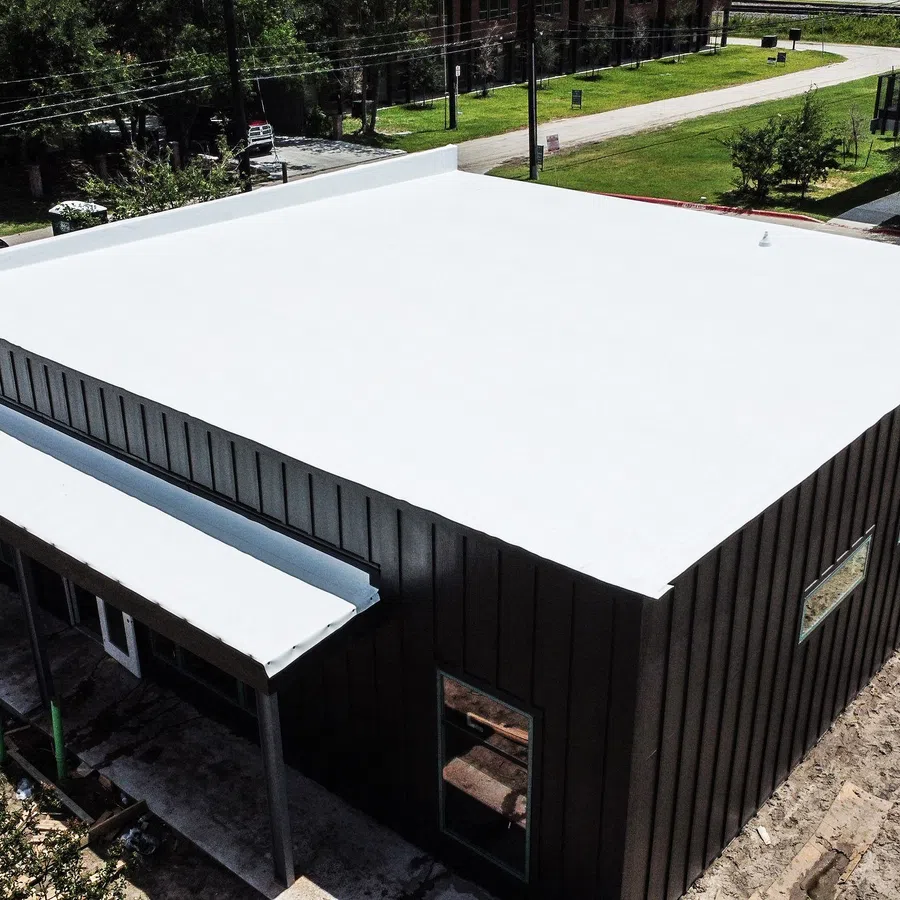 Aerial view of a completed new commercial building with a white flat membrane roof and dark standing seam metal facade, showing the finished roof system and covered entry canopy on a construction site in Texas.