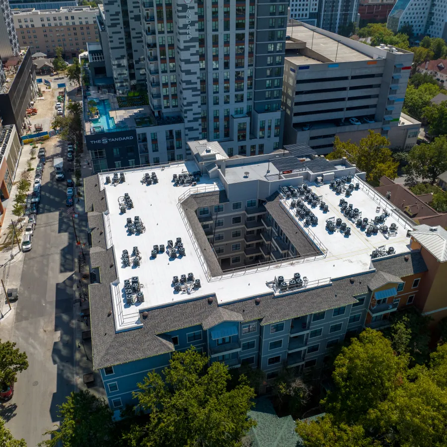 Aerial view of a completed TPO reroof on a student housing community near downtown Austin, Texas, showing the white membrane roof worked around rooftop HVAC units and an interior courtyard, with The Standard high-rise and the city skyline in the background.