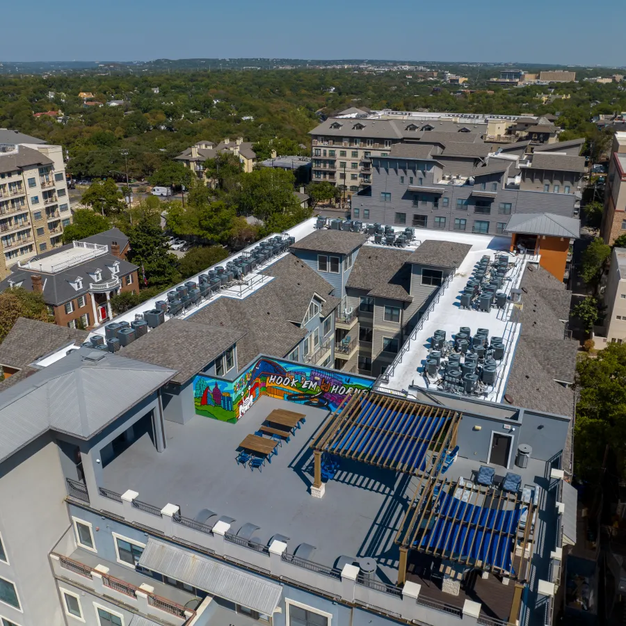 Aerial view of a student housing property in Austin, Texas, showing a rooftop amenity deck with pergola seating and a Hook 'Em Horns mural alongside extensive white TPO membrane roofing and rows of rooftop HVAC units.