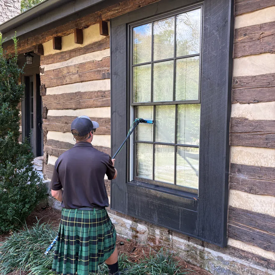 Man in a green plaid kilt cleaning a large window on a rustic log cabin with a window cleaning tool.