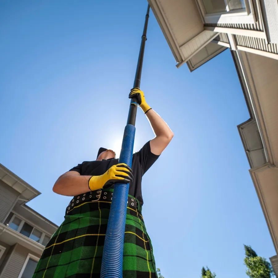 Man in a green kilt using a long pole tool to clean gutters under a clear blue sky.