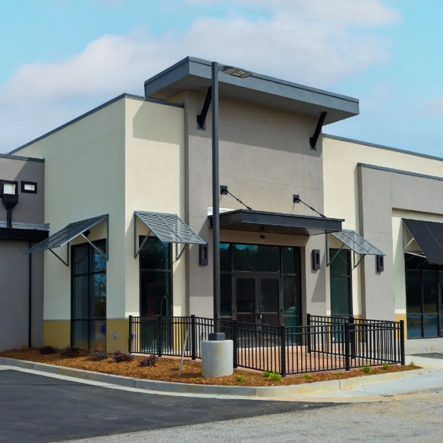 Modern commercial building with large windows, metal awnings, beige and gray exterior, and black railing on a sunny day.