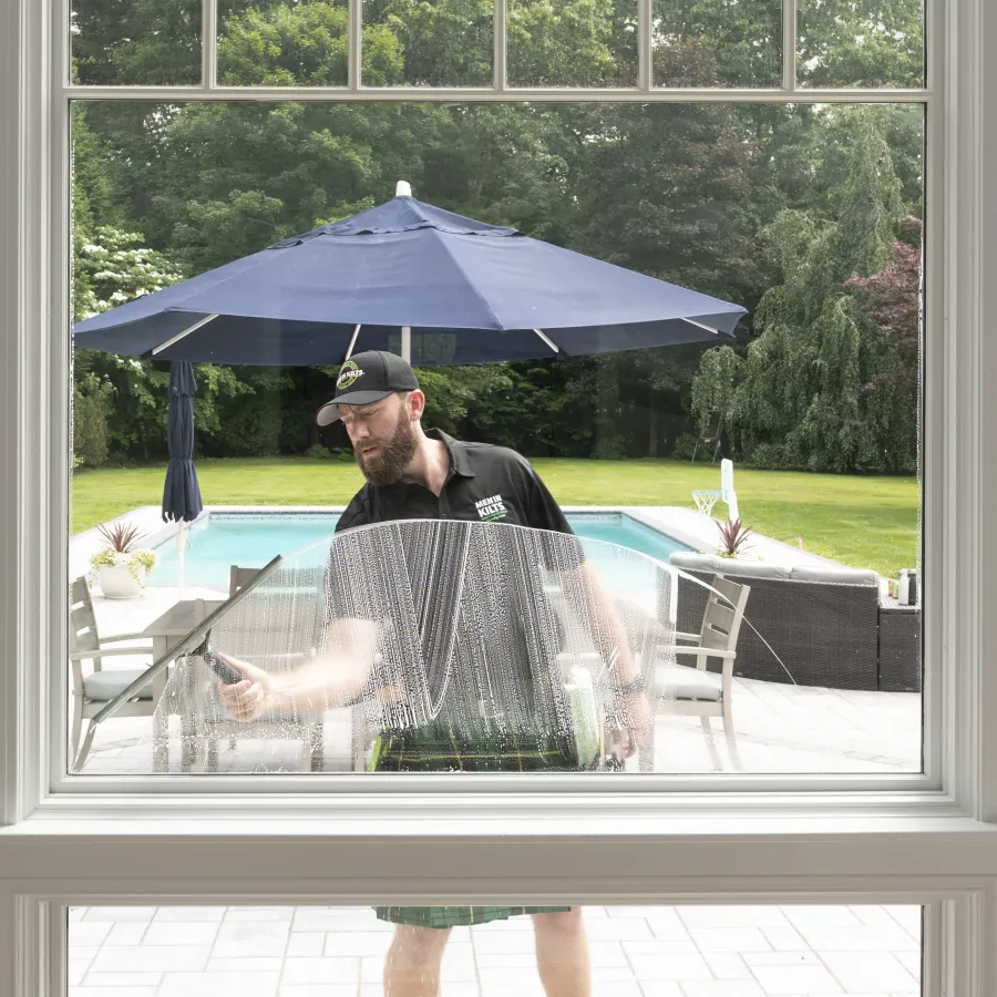 Man cleaning a large window outside near a pool with patio furniture and a blue umbrella in the background