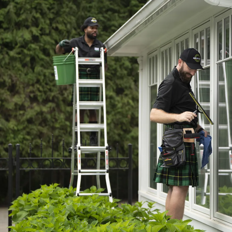 Two professional cleaners wearing kilts cleaning house windows outdoors with tools and ladder near green bushes.