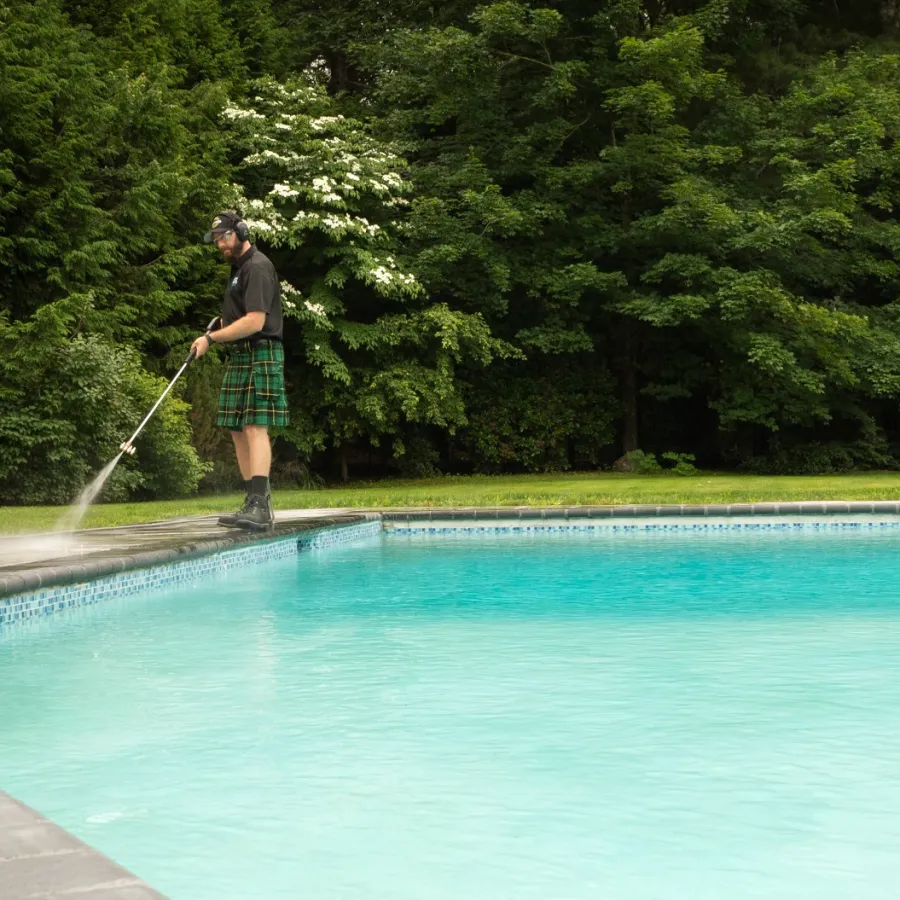 Man in a kilt power washing a patio beside a clear blue swimming pool with dense green trees in the background