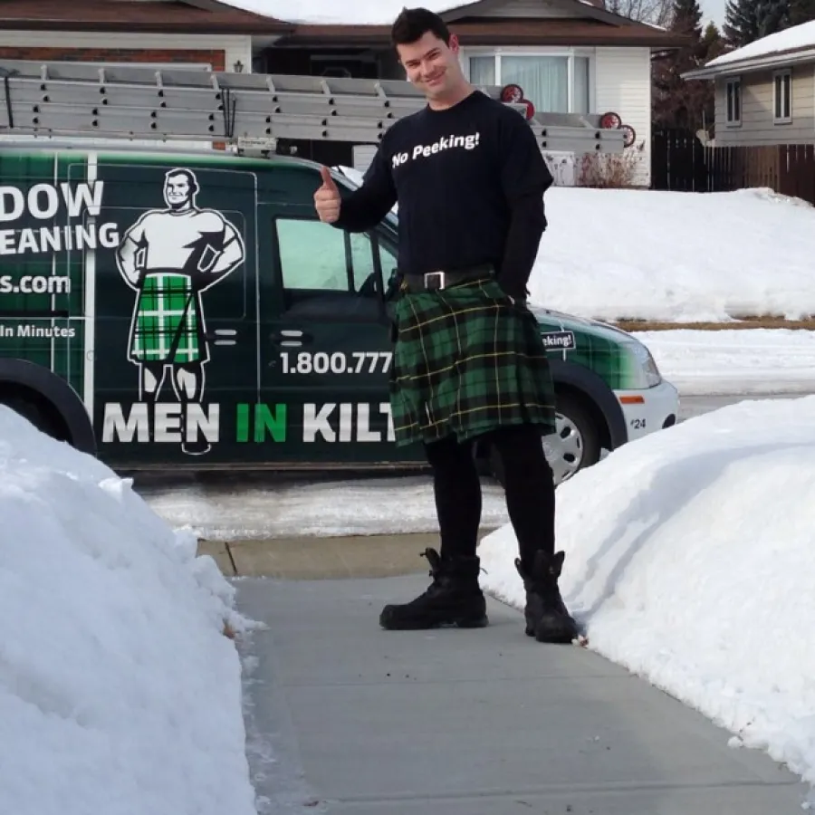 Man in a kilt standing on a snowy sidewalk giving thumbs up next to Men in Kilts window cleaning van.
