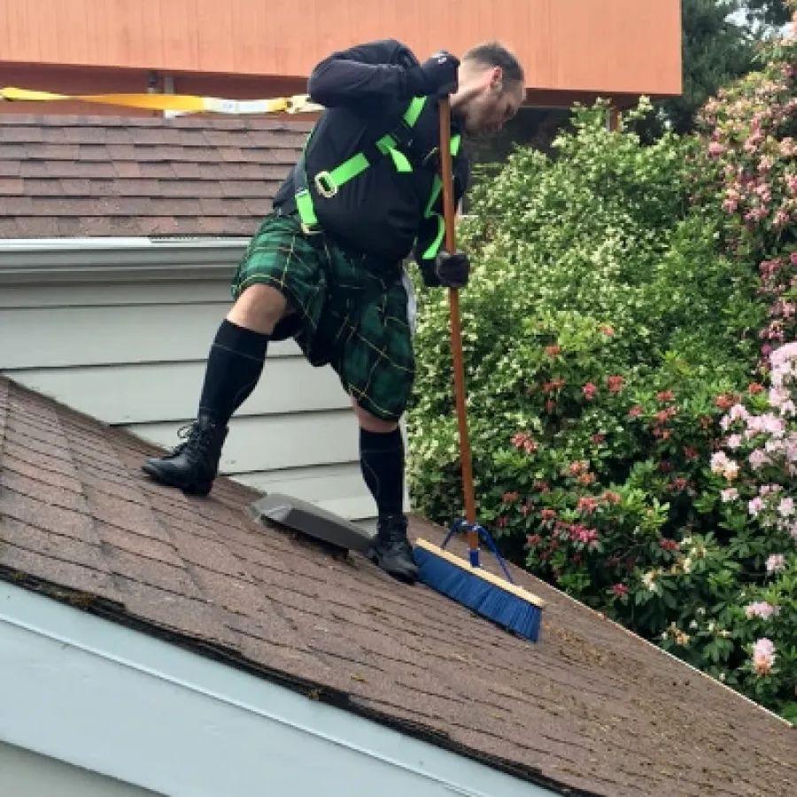 Man wearing a green kilt and safety harness sweeping moss off a sloped roof with a blue brush near flowering bushes.
