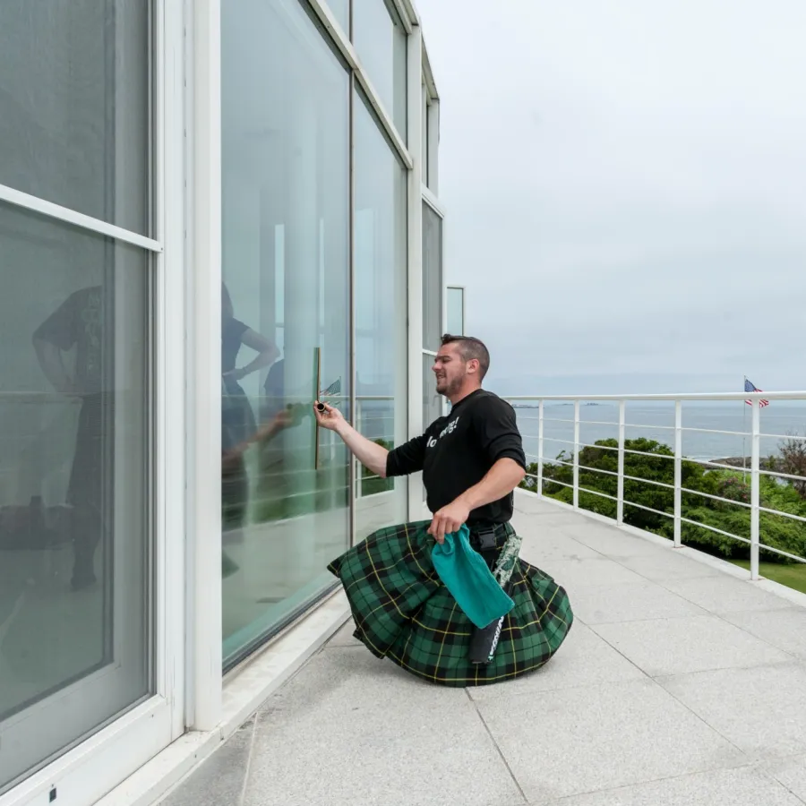 Man wearing a green kilt cleans large glass windows on a modern balcony overlooking the ocean.