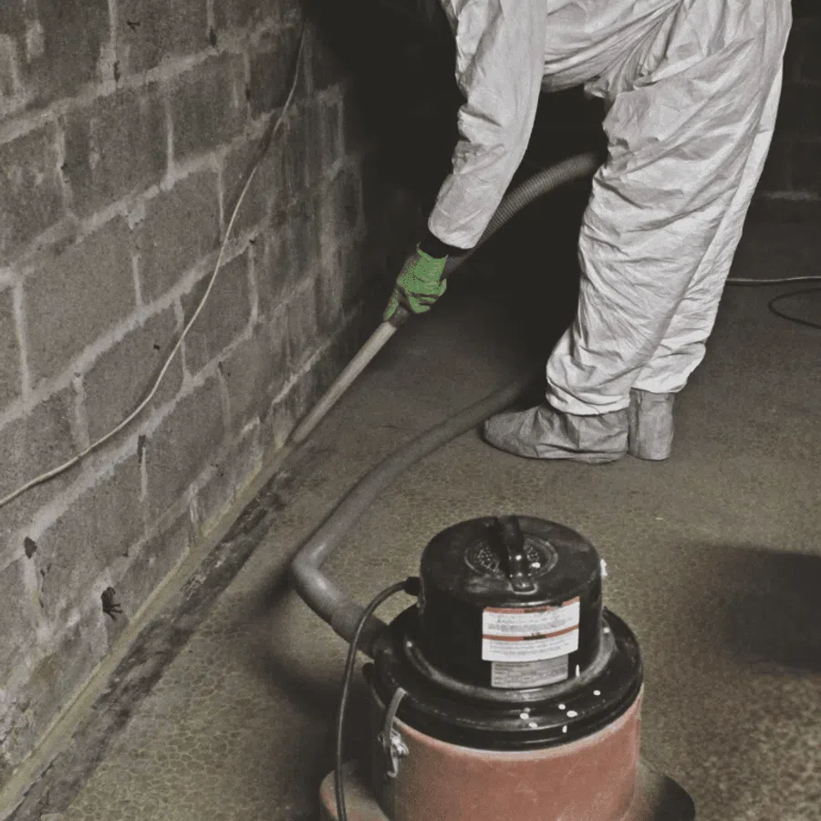 Person in protective suit and gloves vacuuming dust and debris from basement floor near concrete wall.