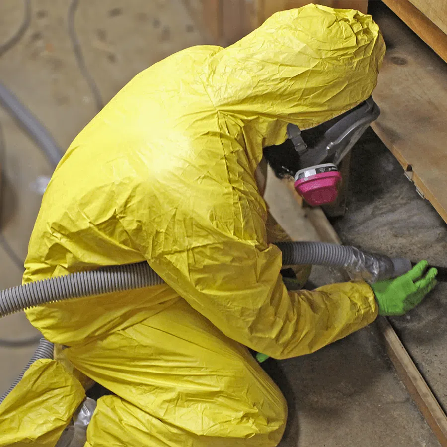 Person in a yellow hazmat suit vacuuming dust from a wooden cabinet using protective gear indoors.