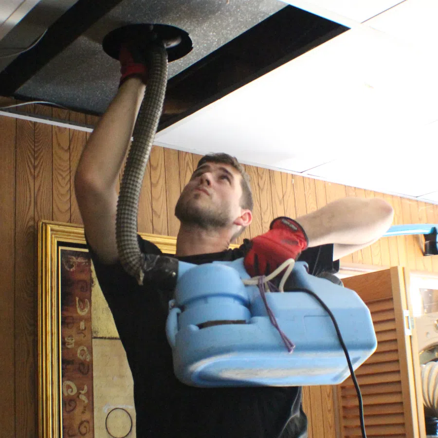 Technician using a blue machine to clean a ceiling vent in a wood-paneled room wearing red gloves.