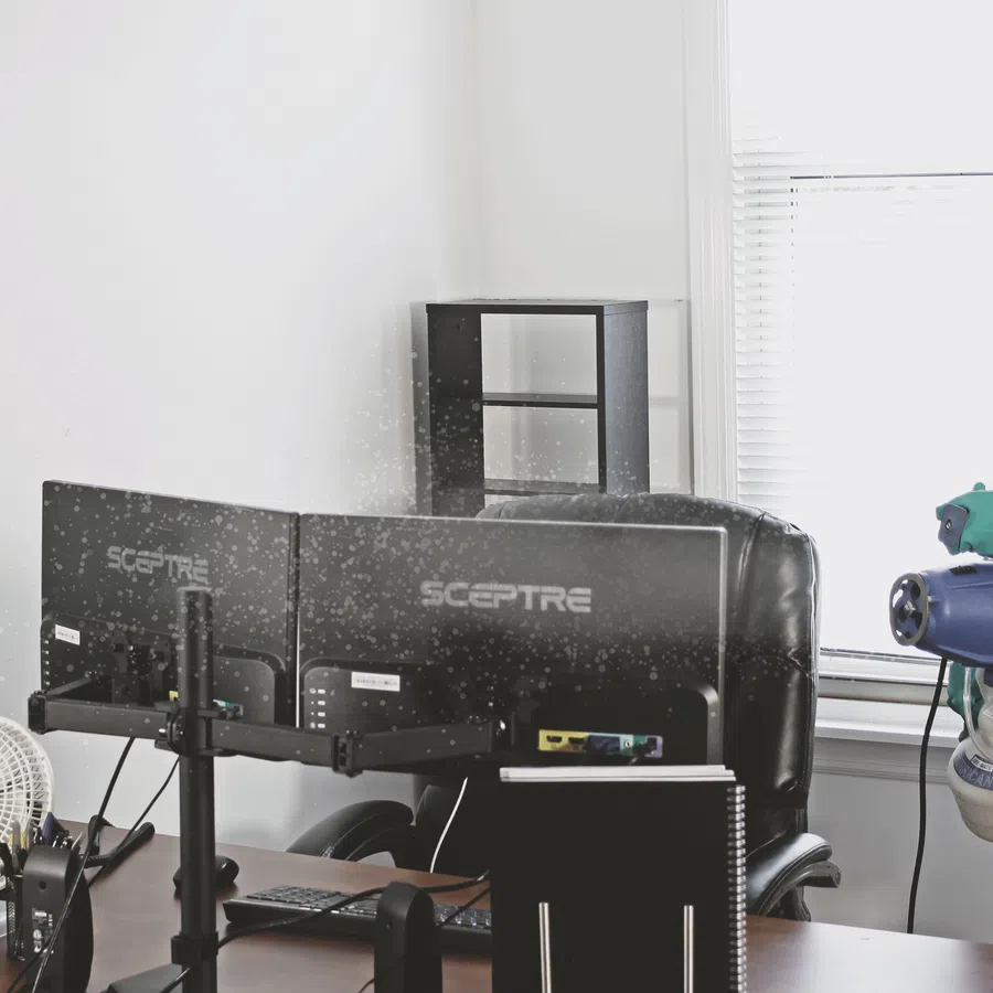 Person in protective suit disinfecting office with dual monitors and desktop equipment.