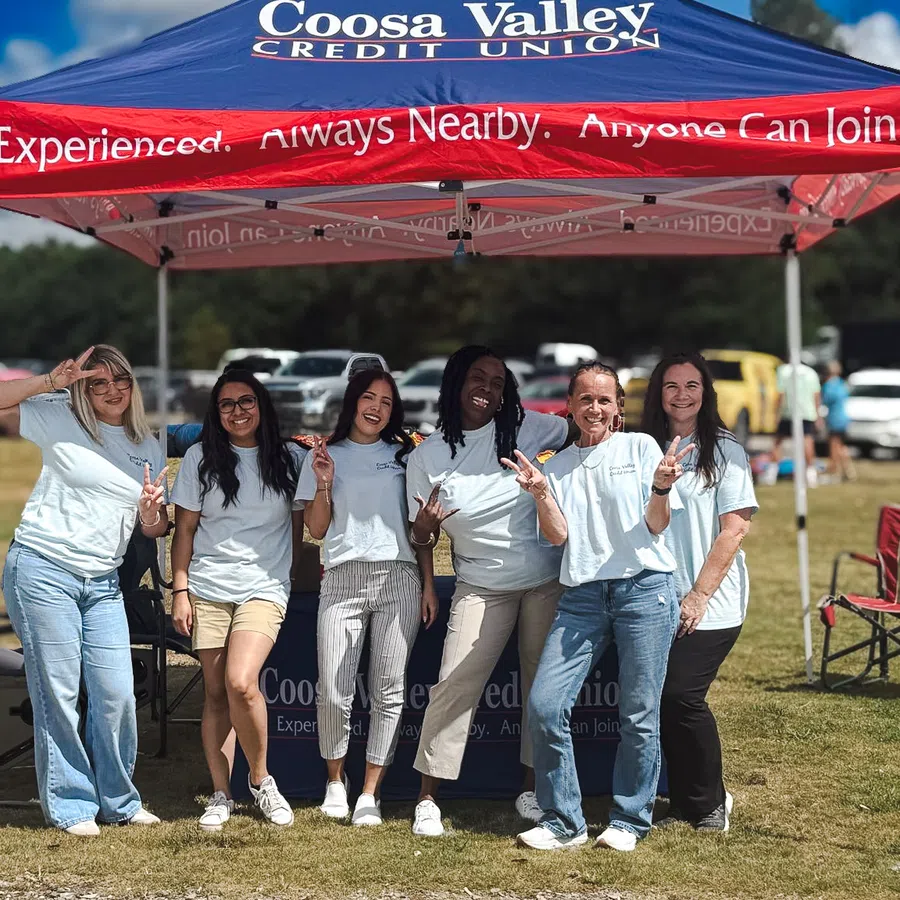 Group of six women posing and smiling under a Coosa Valley Credit Union event tent outdoors on a sunny day.