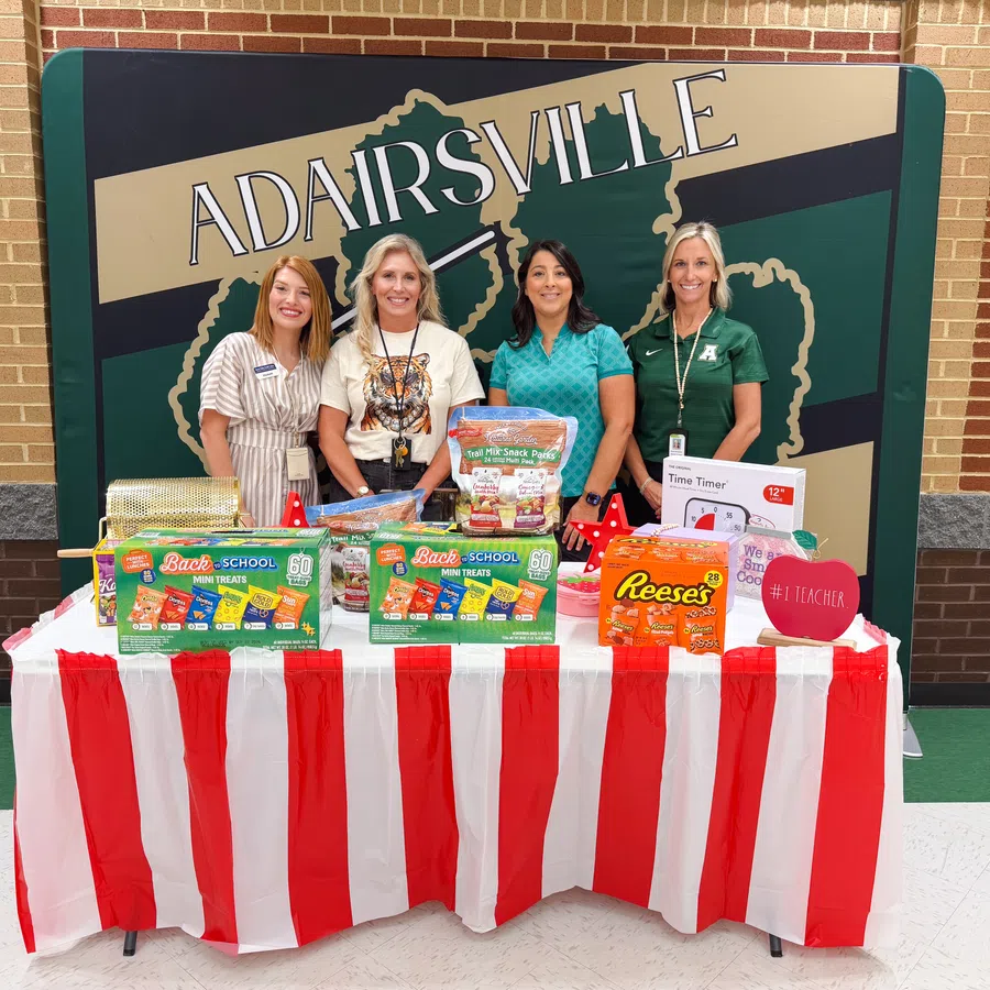 Four women standing behind a table with school supplies and snacks at Adairsville event with red and white striped tablecloth.