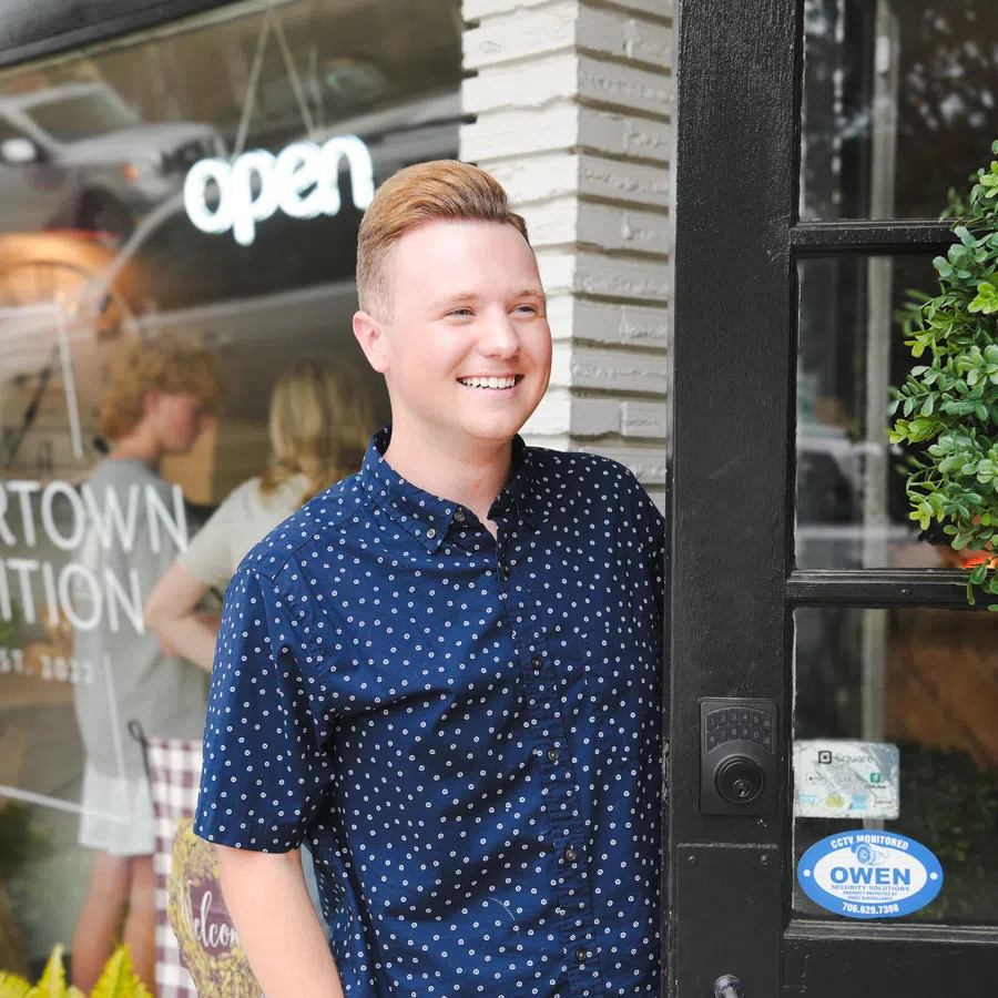 Smiling man stands at the entrance of Cedartown Nutrition store with open sign and greenery outside