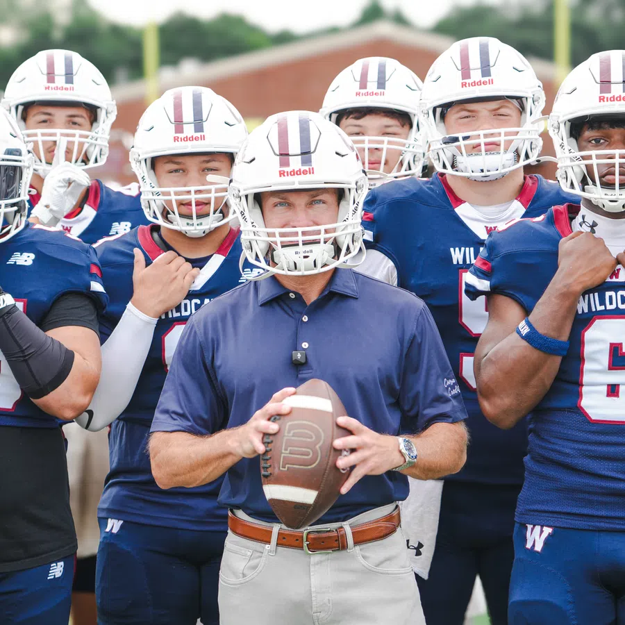 Youth football coach holding ball surrounded by team in Wildcats uniforms and helmets on field.