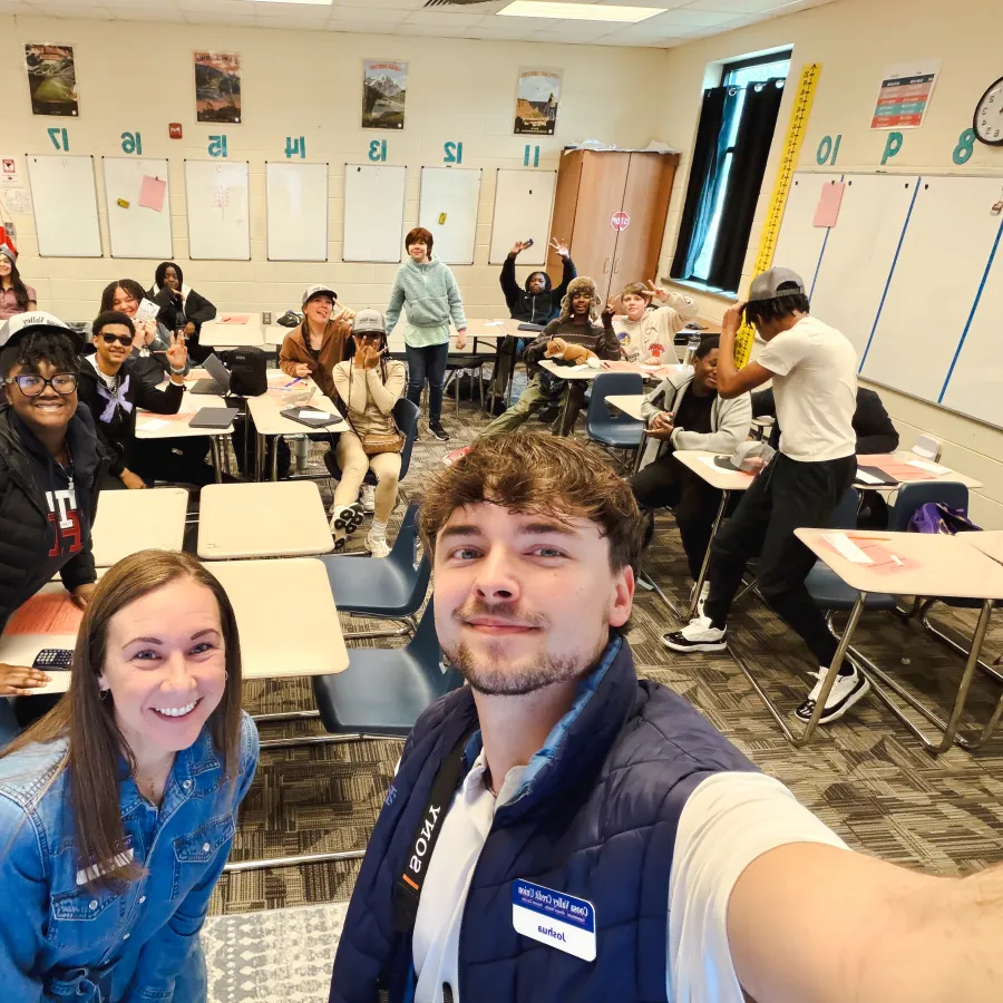 Diverse high school students and teachers smiling together in a bright classroom with desks and educational posters.