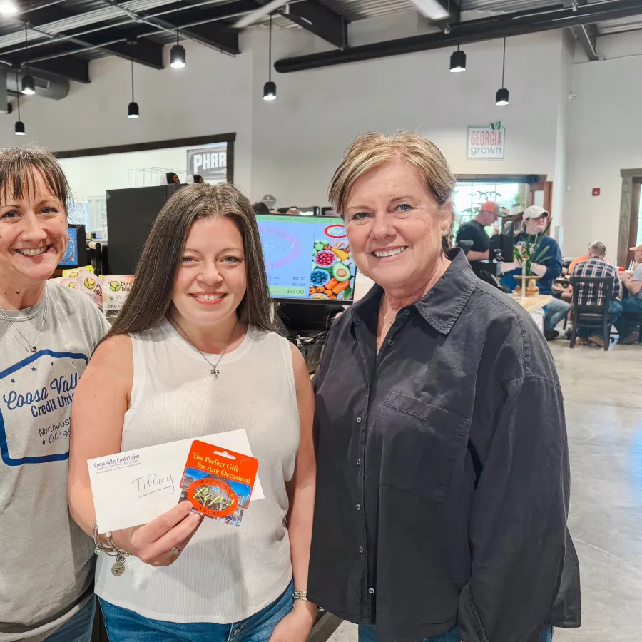 Three women smiling inside a modern store holding an envelope and a gift card with customers in the background