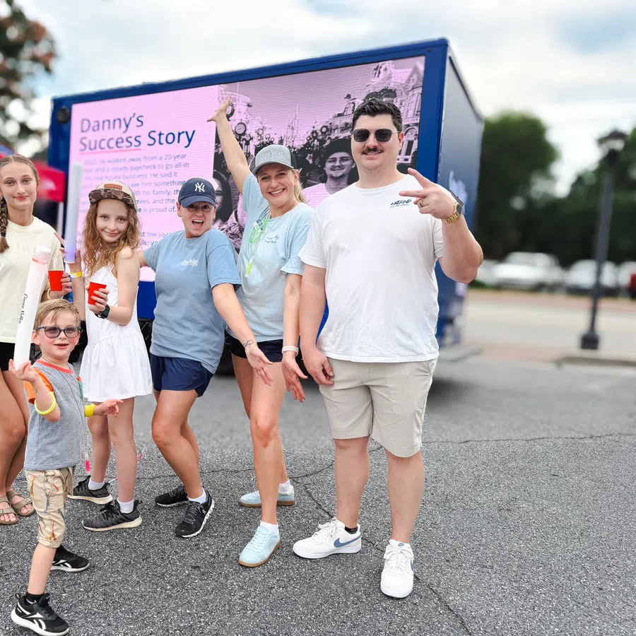 Group of six smiling people posing outdoors near a colorful event banner on a sunny day.