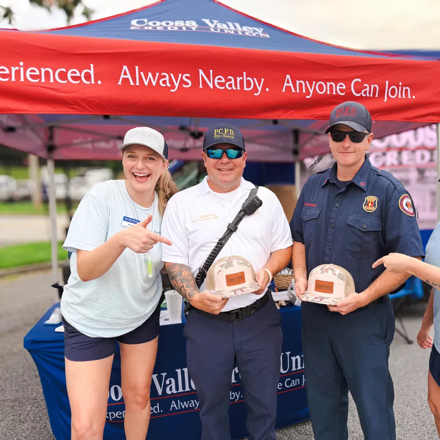 Four people, including two firefighters holding helmets, smiling under a red Coosa Valley Credit Union tent at an outdoor event.