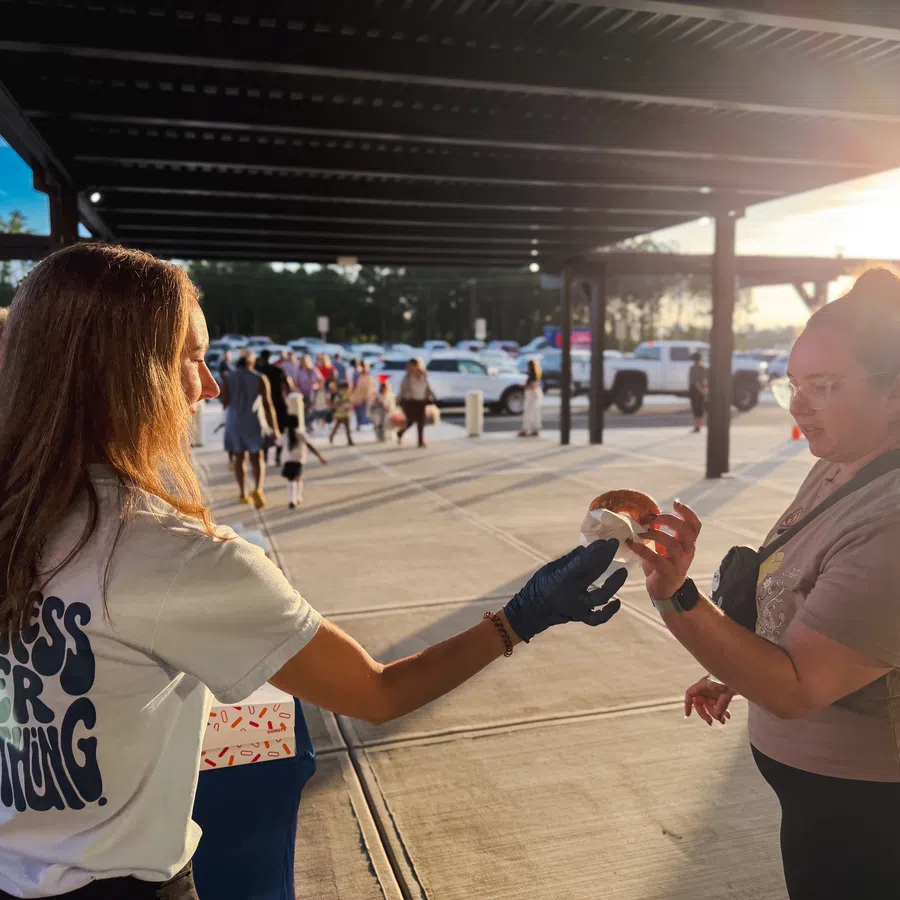 Volunteer handing a donut to a woman under a shaded outdoor area during sunset with parked cars in the background
