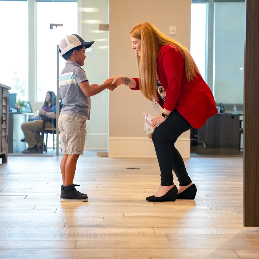 Woman in red jacket smiling and shaking hands with young boy wearing a cap in a bright room