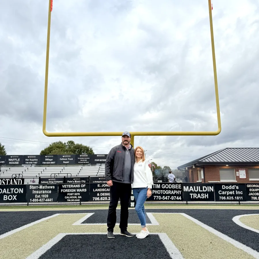 Man and woman standing together on a football field end zone under the goalpost on a cloudy day.