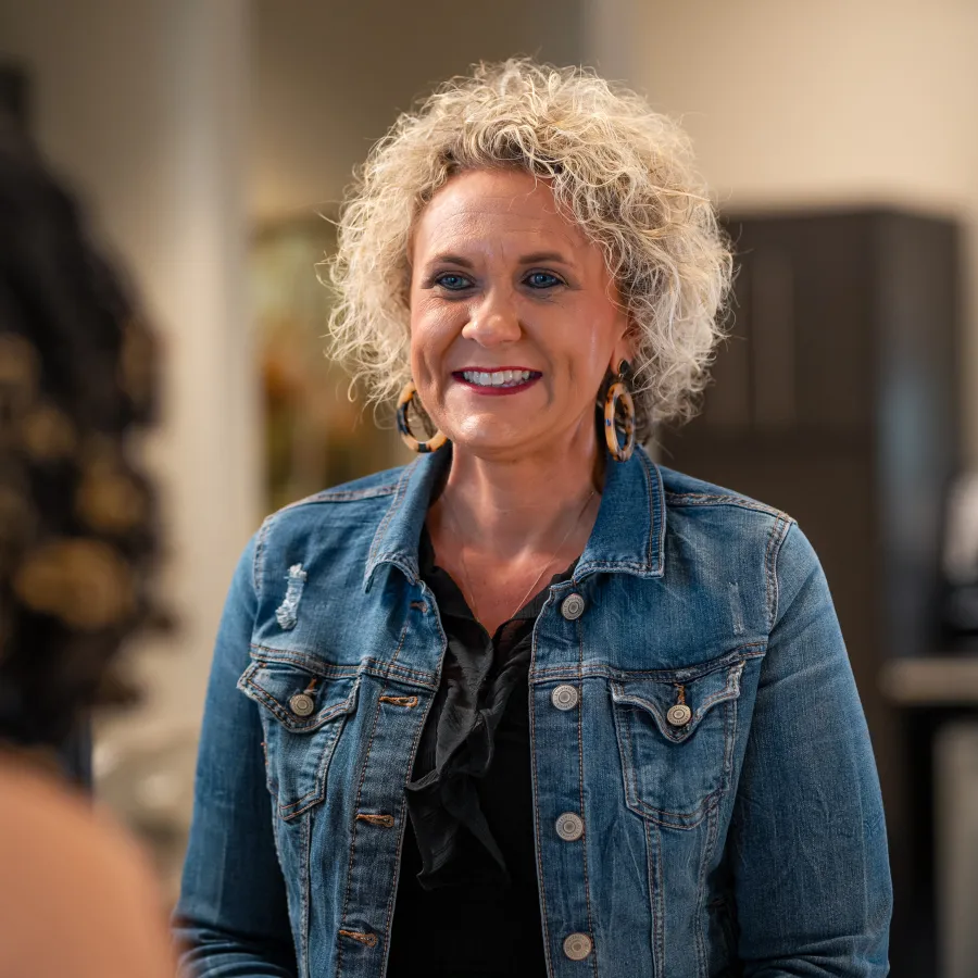 Smiling woman with curly hair in denim jacket talking to another person indoors in a casual setting