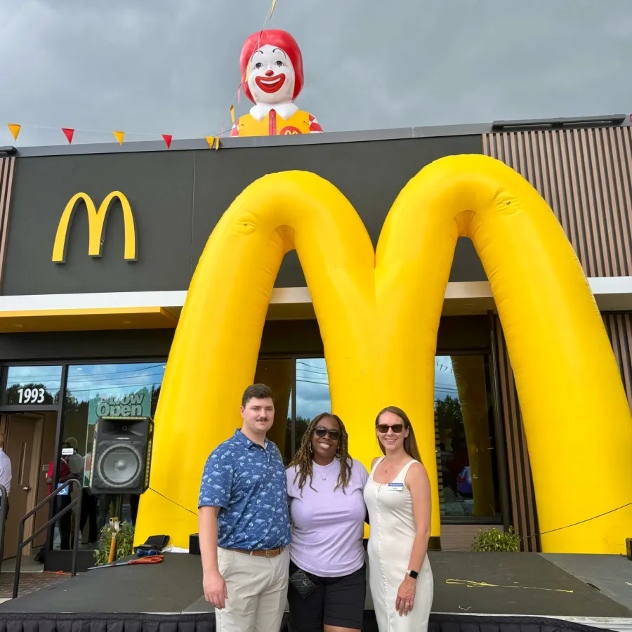 Three people posing in front of a McDonald's restaurant with a large inflatable golden arches and Ronald McDonald figure