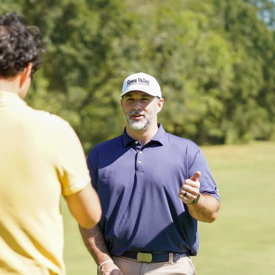 Two men on golf course, one in yellow shirt with back to camera, the other in navy shirt and white cap speaking.