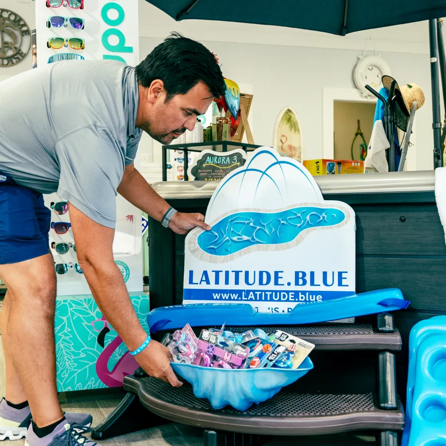 Man arranging Latitude.Blue branded pool accessories and sunglasses inside a retail store near hot tub display.
