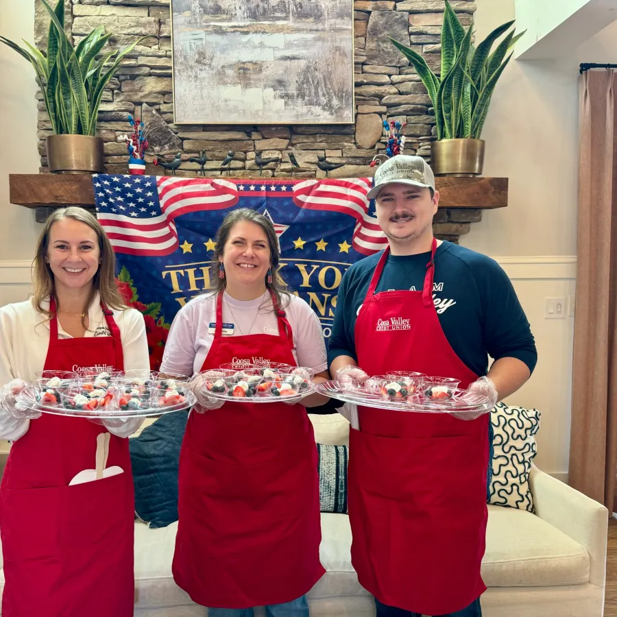 Three people wearing red aprons holding trays of cupcakes in front of a patriotic banner and stone wall.