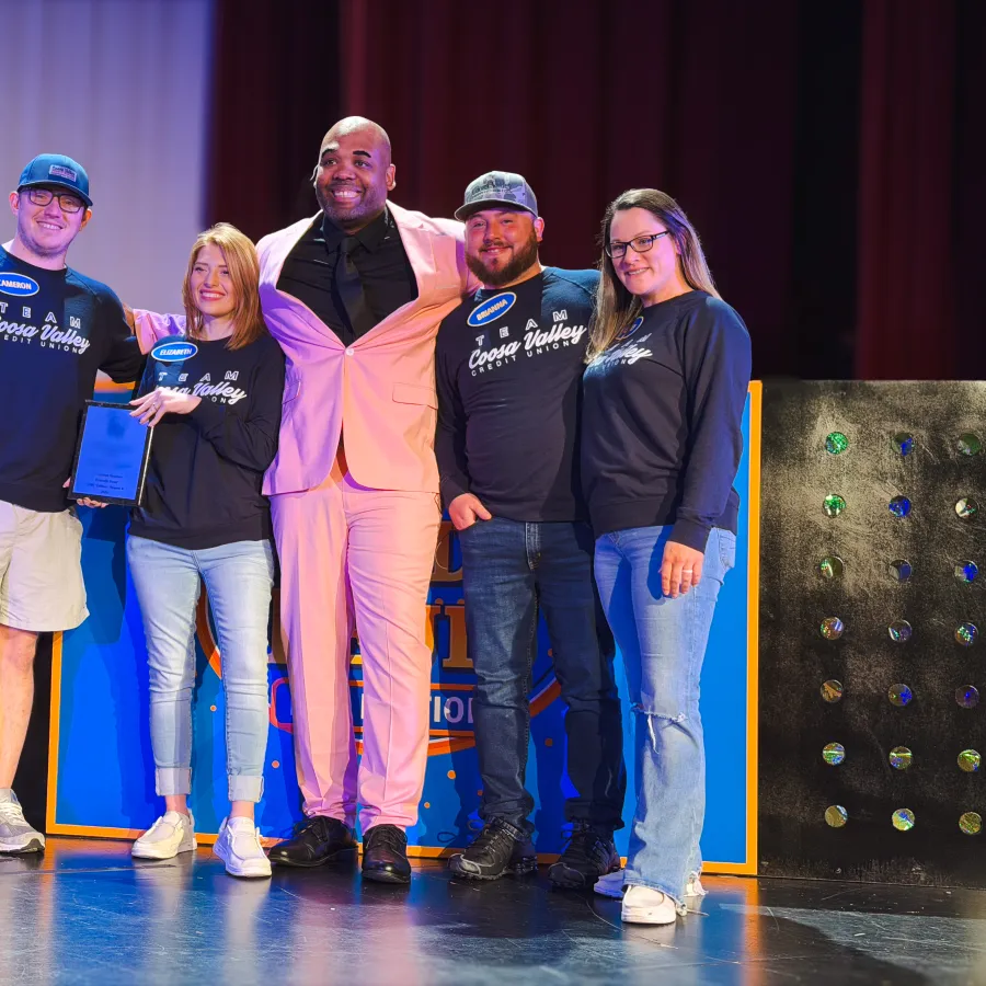 Group of five people smiling on stage, three wearing Coosa Valley logos, one in a peach suit holding an award.