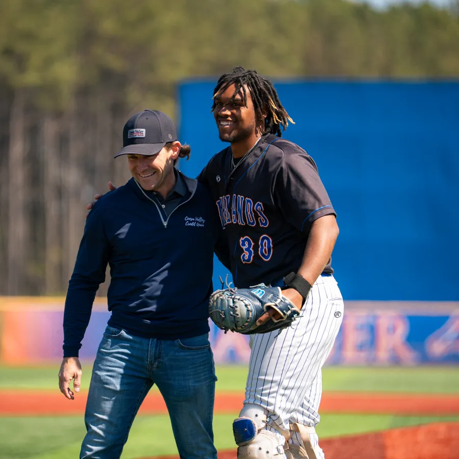 Two men smiling and embracing on a sunny baseball field with one in uniform and the other in casual wear.