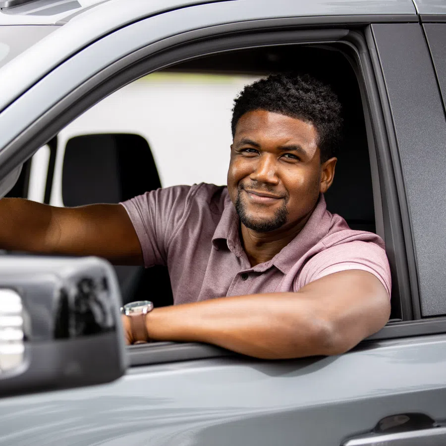 Smiling man in a purple shirt sitting in the driver's seat of a gray car with his arm resting on the window.