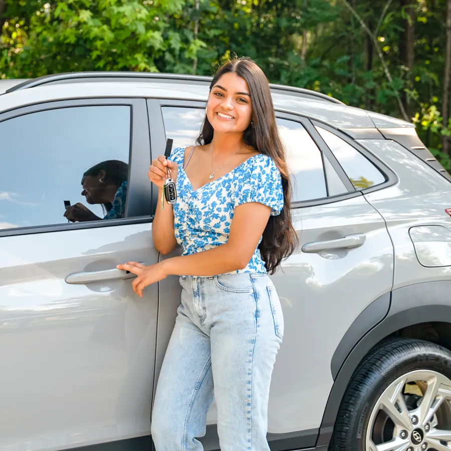 Smiling young woman holding car keys while standing next to a silver SUV with trees in background