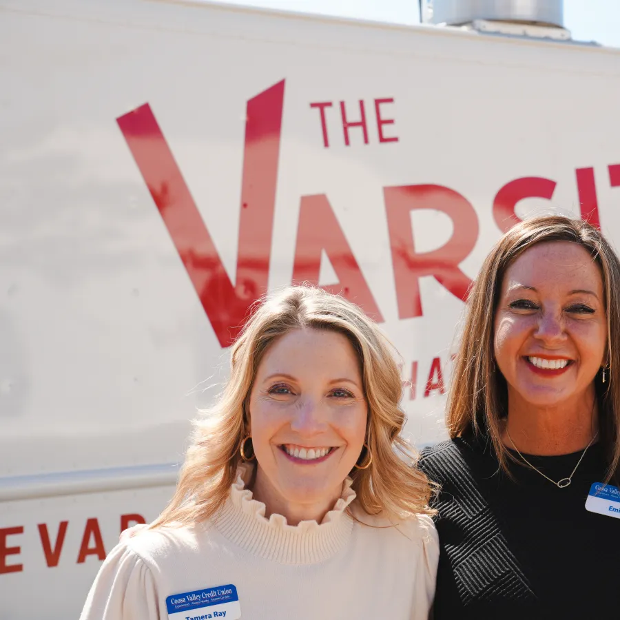 Two smiling women with name tags standing in front of The Varsity food truck on a sunny day