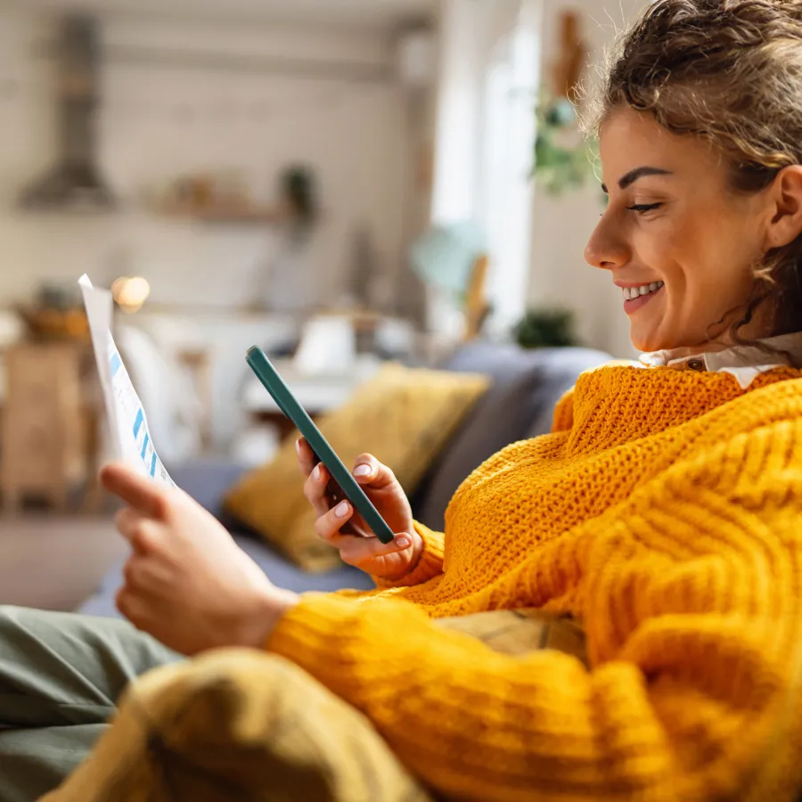 Smiling woman in a mustard sweater using smartphone while holding paper on a cozy living room couch.
