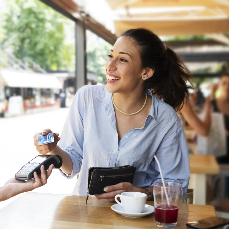Smiling woman paying with credit card at outdoor café while holding wallet, with drinks on the table.