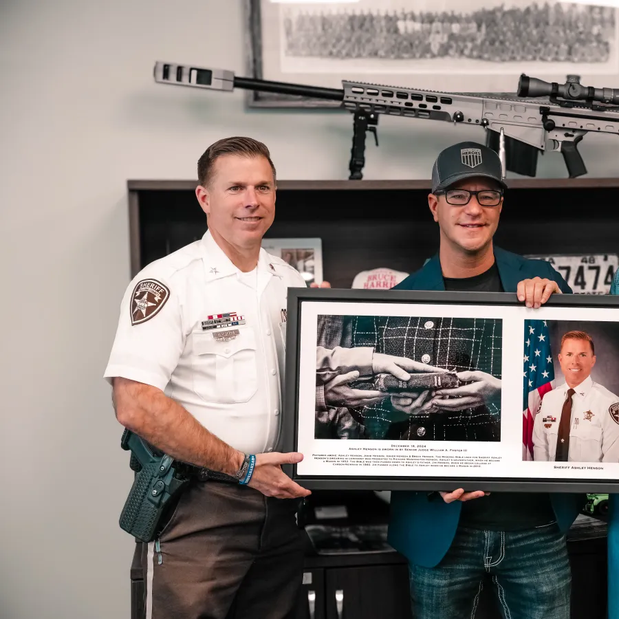 Three people holding a framed tribute photo of a police officer with a rifle displayed on the shelf behind them